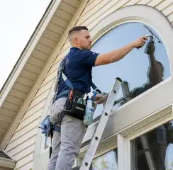 Professional window cleaner on a ladder using a squeegee to clean an exterior arched window, Syracuse, NY.
